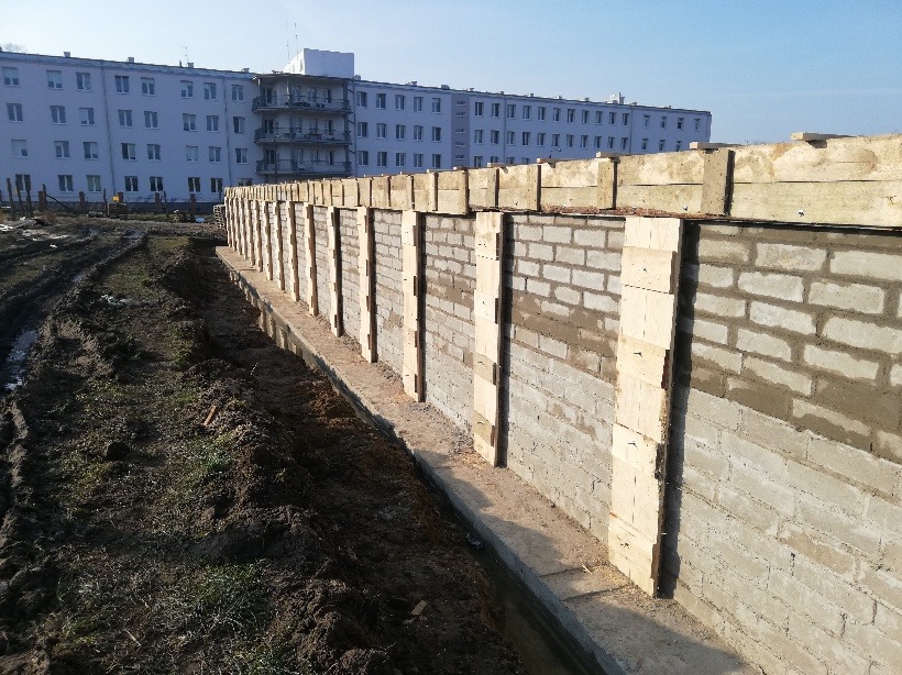 Wznoszenie ściany fundamentowej z bloczków betonowych, wspartej drewnianymi stemplami, widoczne świeże ślady zaprawy, w tle budynek wielorodzinny.