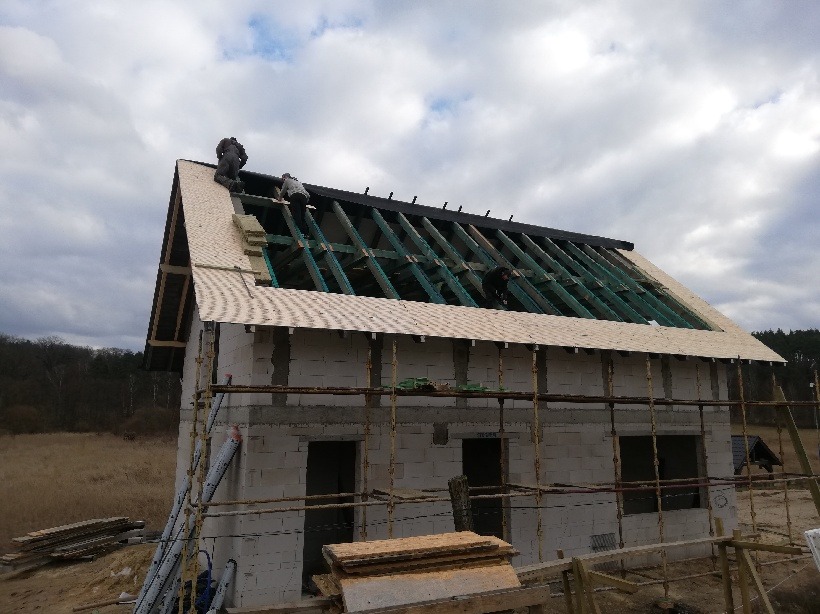 Budowa domu jednorodzinnego: murowana konstrukcja w stanie surowym otwartym z widocznym szkieletem dachu i pracownikami montującymi elementy poszycia dachowego.