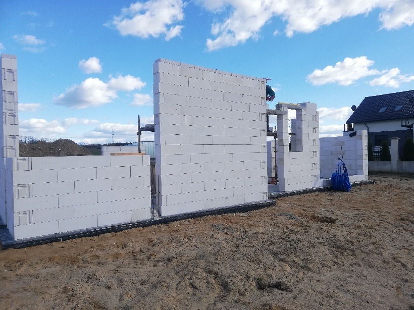 Wznoszone ściany parteru domu z betonu komórkowego, widoczny murarz przy pracy, w tle nowy dom jednorodzinny i błękitne niebo.