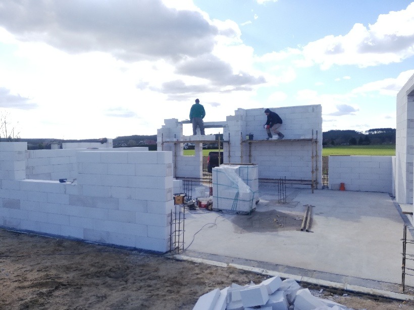 Wznoszenie ścian domu z białych bloczków na fundamencie, dwóch pracowników na rusztowaniach, widok na teren budowy i odległe pola w słoneczny dzień.