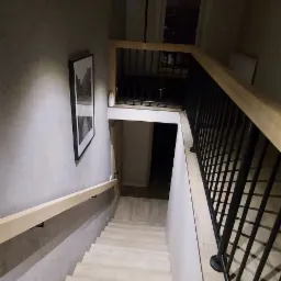 Nowoczesne schody z drewnianymi stopniami i czarną metalową balustradą, oświetlone minimalistyczną lampą wiszącą, z obrazem na ścianie i widokiem na dolny poziom.