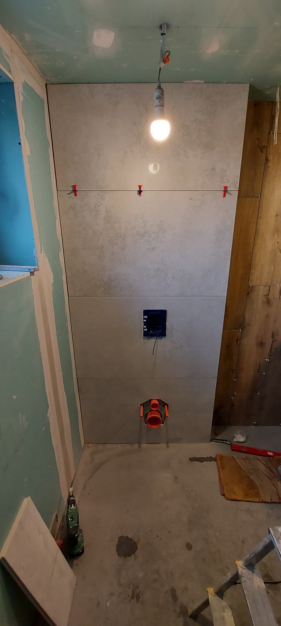 Montaż płytek ceramicznych na ścianie w łazience, widoczne krzyżyki dystansowe, instalacja podtynkowa WC, surowe ściany z płyt gipsowo-kartonowych i drewnianych paneli.