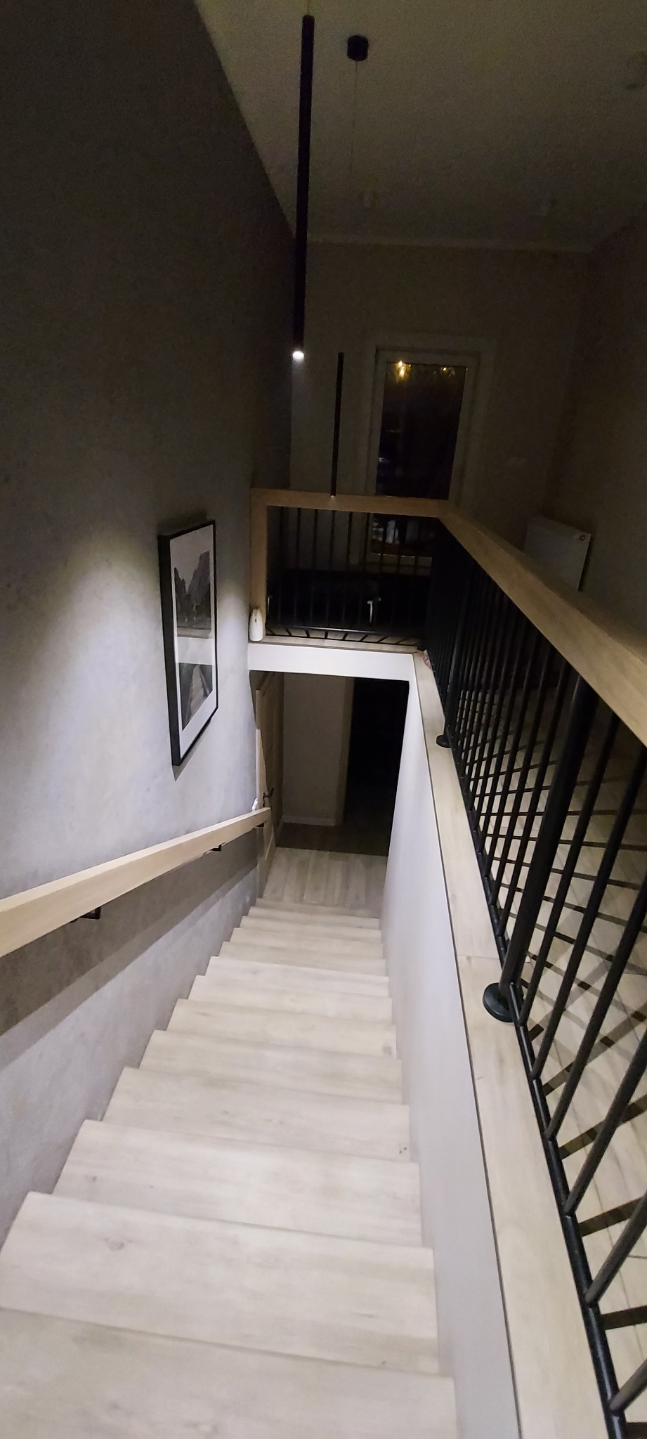 Nowoczesne schody z drewnianymi stopniami i czarną metalową balustradą, oświetlone minimalistyczną lampą wiszącą, z obrazem na ścianie i widokiem na dolny poziom.