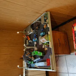 Makieta składu zabawek z miniaturowymi ciężarówkami, koparkami i węglem, oświetlona małymi lampkami, ustawiona w pomieszczeniu z drewnianą boazerią i wagą elektroniczną na ścianie.