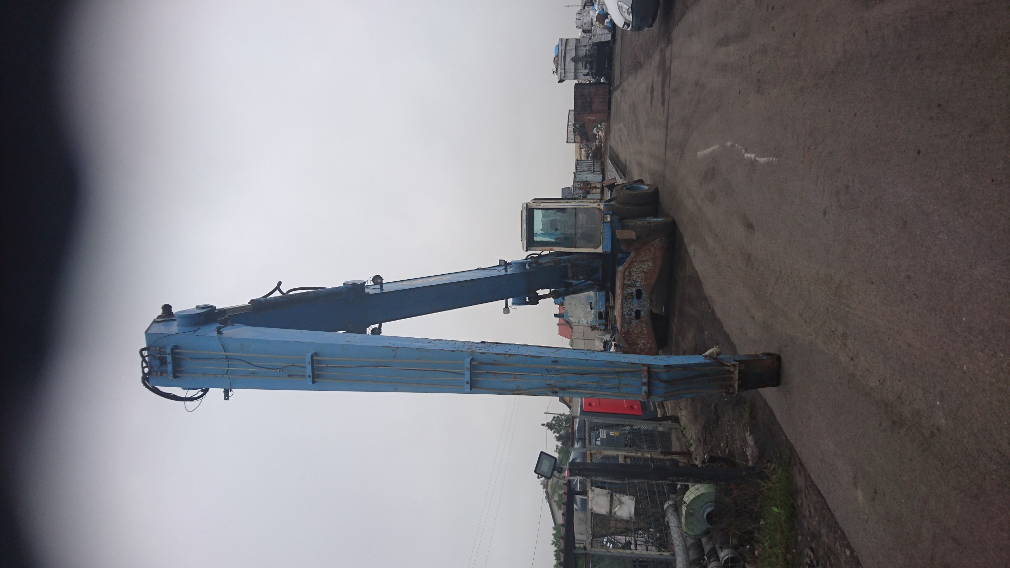 Niebieska, starsza koparka z wysuniętym ramieniem na placu składowym, widoczne inne maszyny i elementy konstrukcyjne w tle.