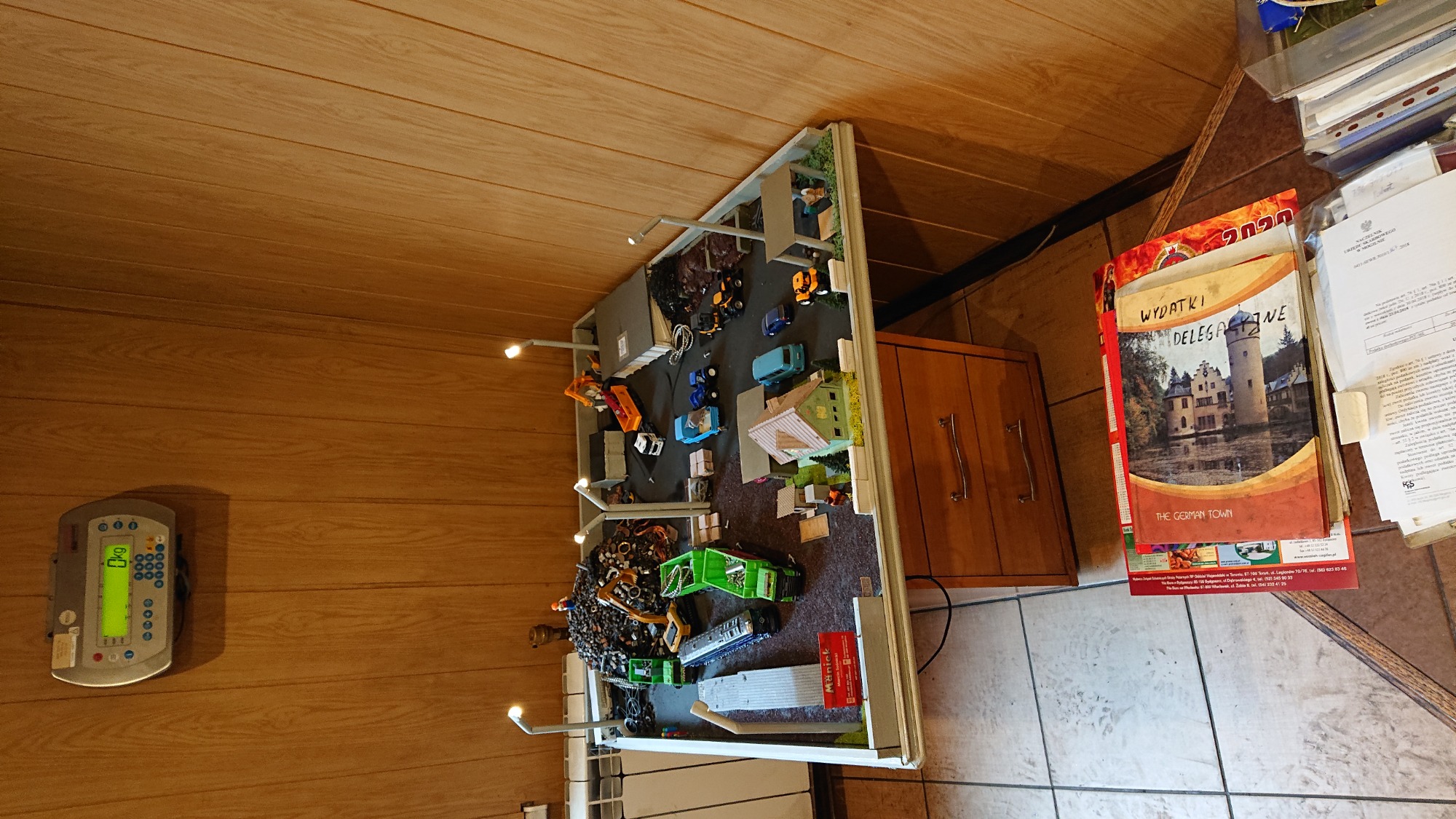 Makieta składu zabawek z miniaturowymi ciężarówkami, koparkami i węglem, oświetlona małymi lampkami, ustawiona w pomieszczeniu z drewnianą boazerią i wagą elektroniczną na ścianie.