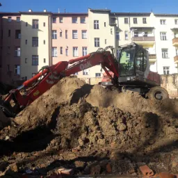 Czerwony koparka O&K pracująca na placu budowy w mieście, z widocznymi budynkami mieszkalnymi w tle, usuwa ziemię.