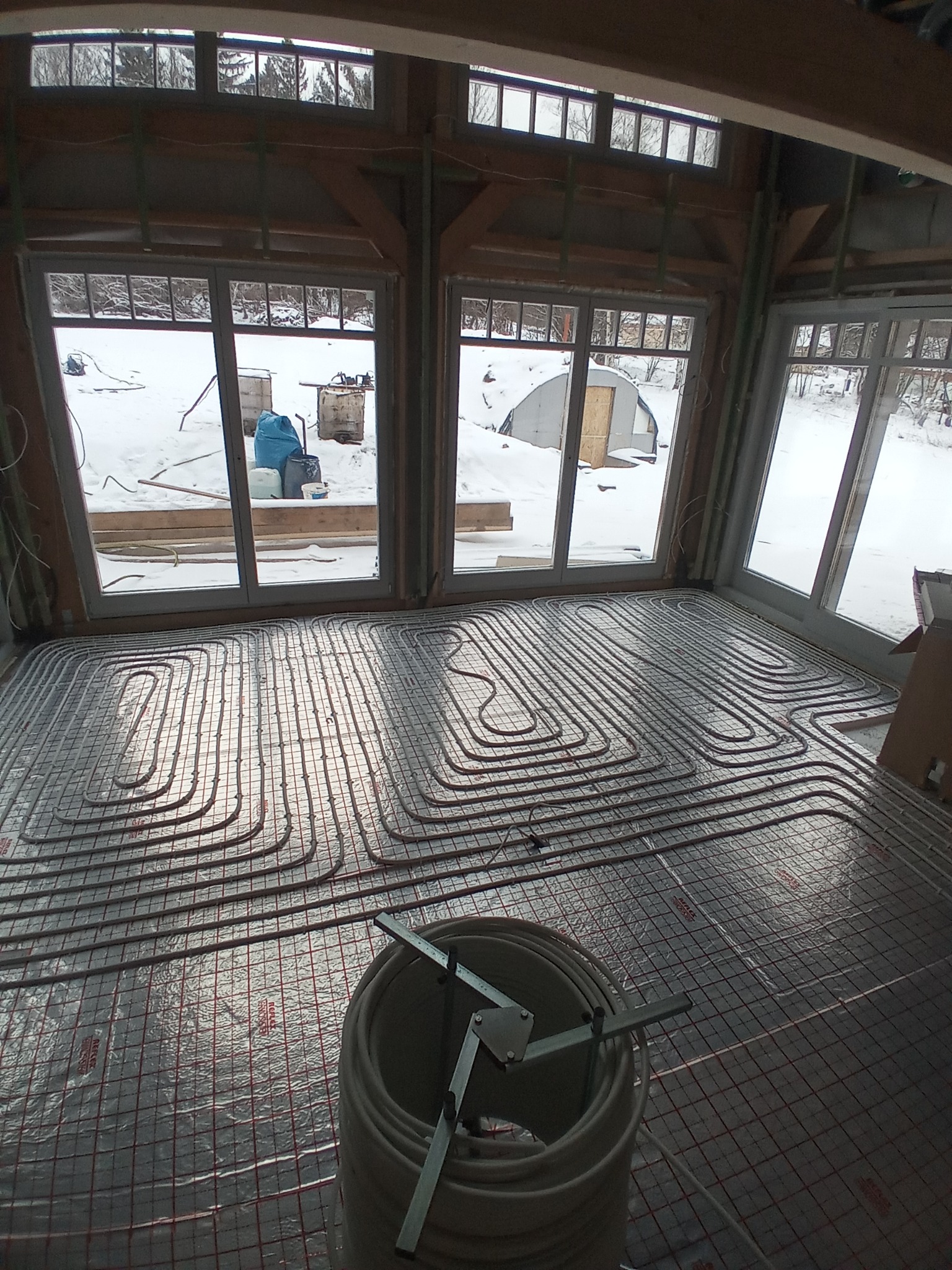 Wnętrze w trakcie budowy z widoczną instalacją ogrzewania podłogowego na folii refleksyjnej z siatką, rolka rur i narzędzia w rogu, za oknami zimowy krajobraz.