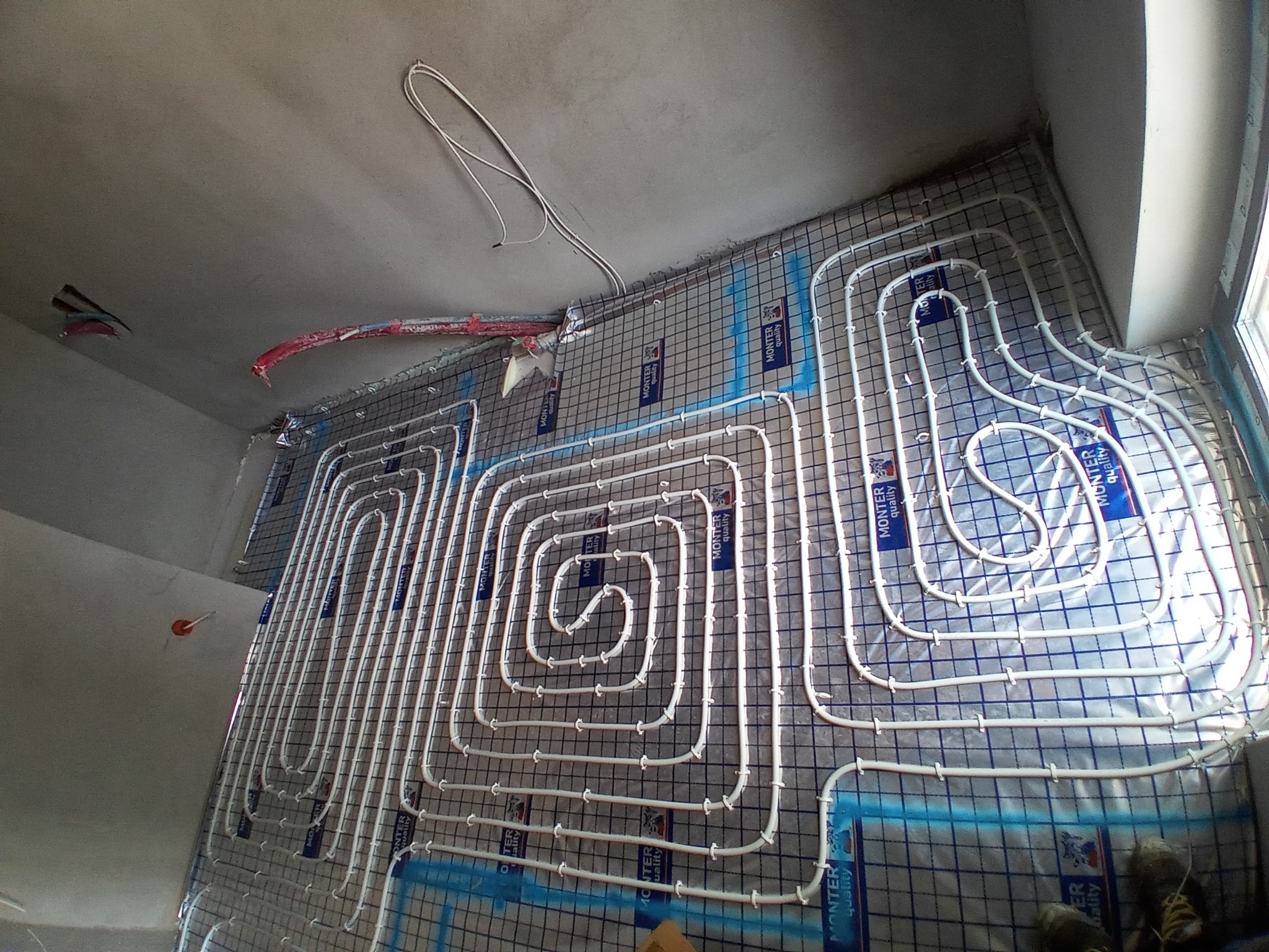Białe rurki instalacji ogrzewania podłogowego ułożone spiralnie na siatce montażowej, widoczne przewody wychodzące ze ściany i fragment okna w rogu pomieszczenia.