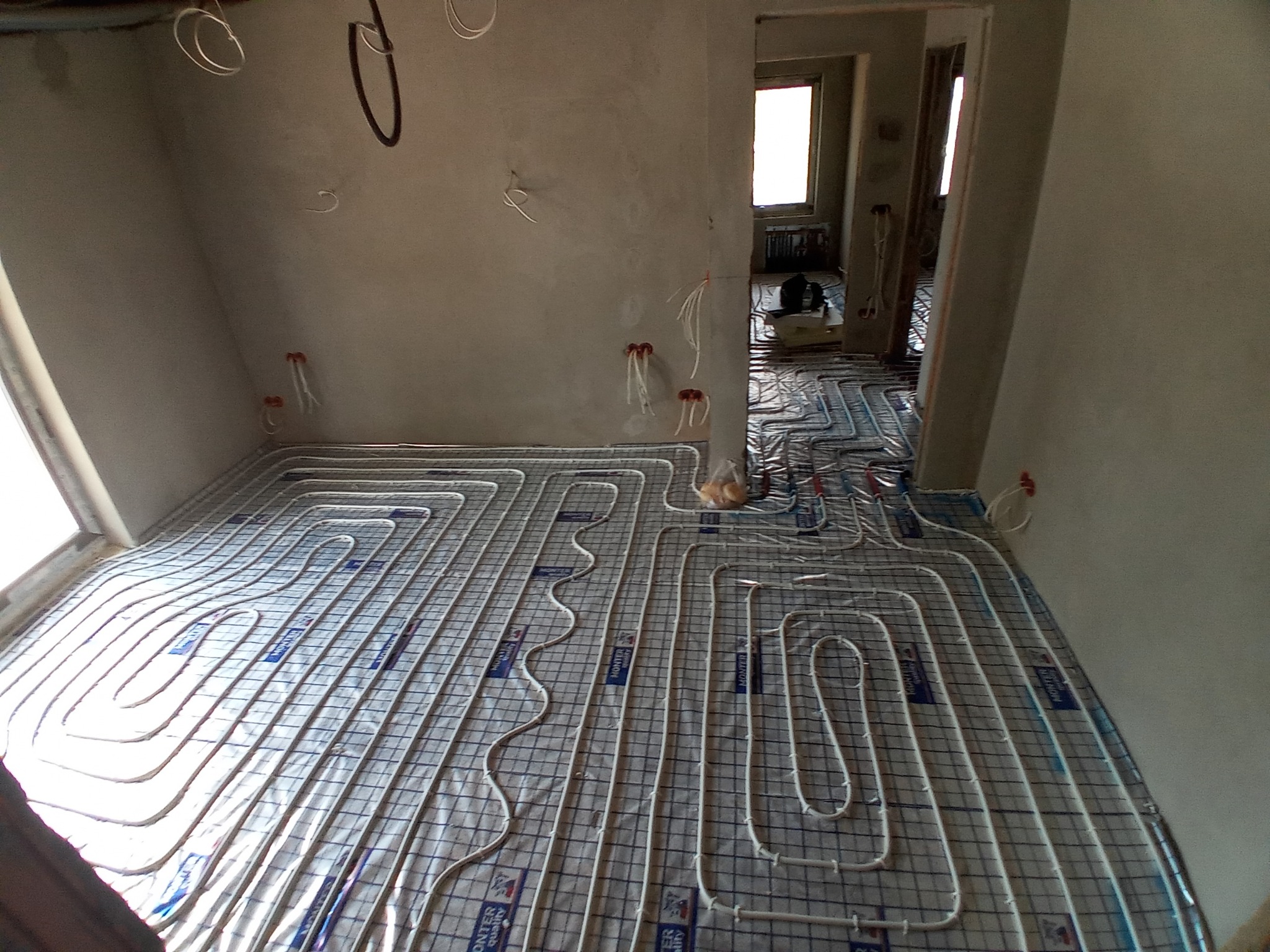 Widok z góry na rozłożoną instalację ogrzewania podłogowego w surowym stanie, rury ułożone na siatce z widocznymi niebieskimi elementami mocującymi, ściany w stanie deweloperskim, w tle widoczne...