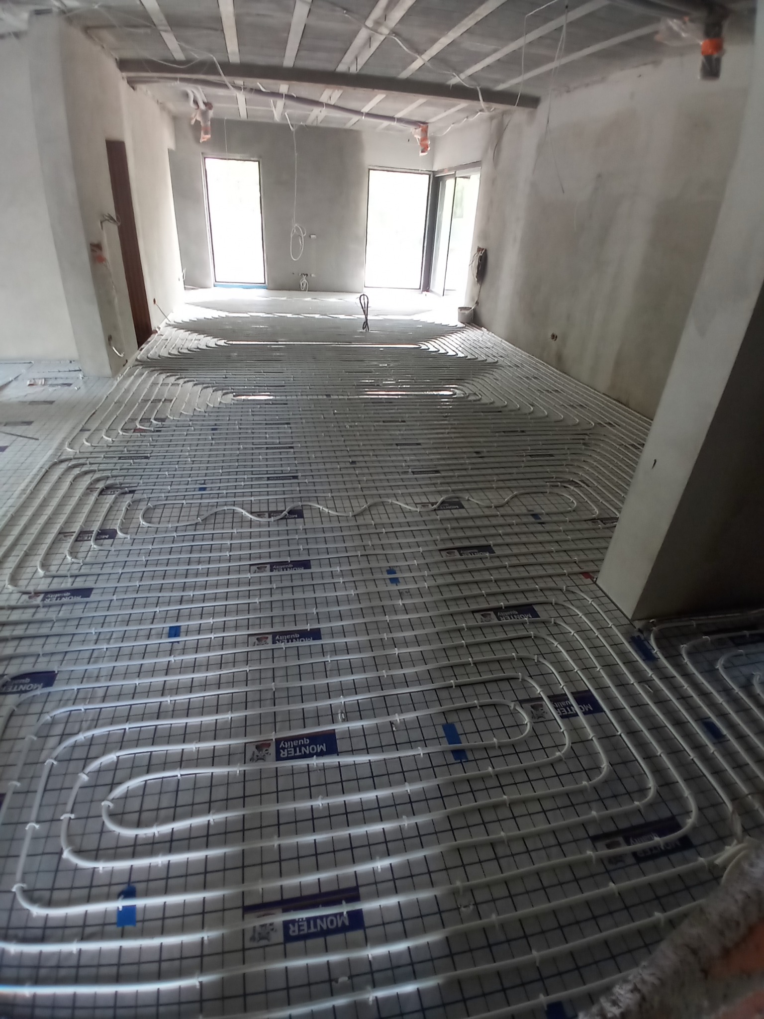 Wnętrze w stanie surowym z widoczną instalacją ogrzewania podłogowego, rurki ułożone w regularnych pętlach na siatce, przygotowane do zalania wylewką.