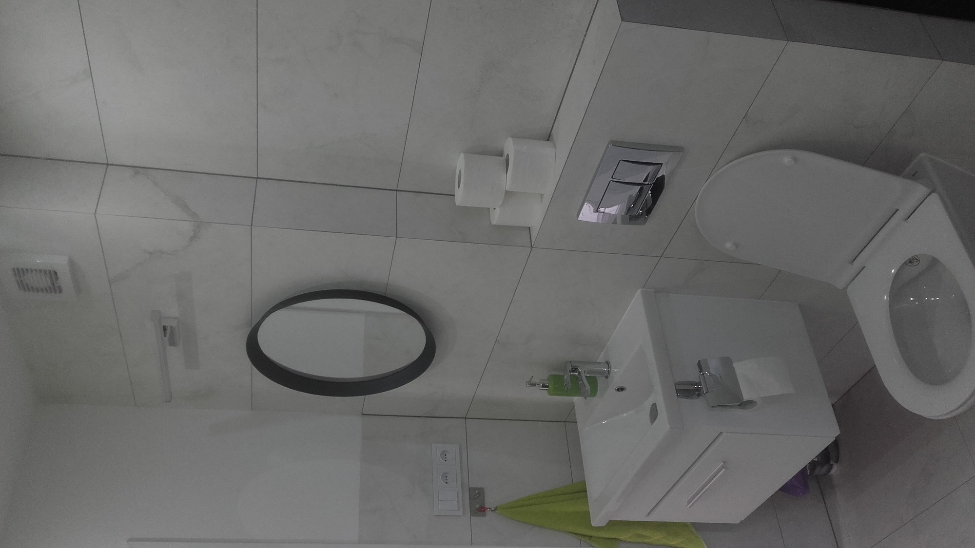 Wykończona łazienka w Potoku Wielkim z białymi płytkami imitującymi marmur, okrągłe lustro w czarnej ramie, podwieszana toaleta i umywalka z szafką, widoczne trzy rolki papieru toaletowego.