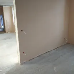 Świeżo otynkowane ściany w surowym pomieszczeniu z wystającymi przewodami elektrycznymi, widok na sąsiednie pomieszczenie z workami i narzędziami.