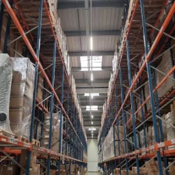 Pełny magazyn z wysokimi regałami wypełnionymi kartonami, oświetlony światłem dziennym z okien dachowych i sztucznym oświetleniem LED.