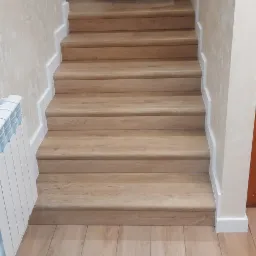 Schody wykończone panelami drewnopodobnymi, z białymi listwami przypodłogowymi i fragmentem grzejnika z niebieskimi akcentami, widok z dołu, w tle osoba w niebieskiej koszulce i szortach.