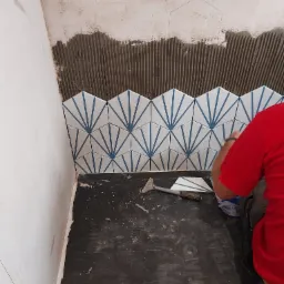 Układanie dekoracyjnych, białych płytek z niebieskimi wzorami na ścianie; widoczny rządek ułożonych płytek, fragment ściany pokryty klejem, narzędzia i osoba w czerwonej koszulce podczas pracy.