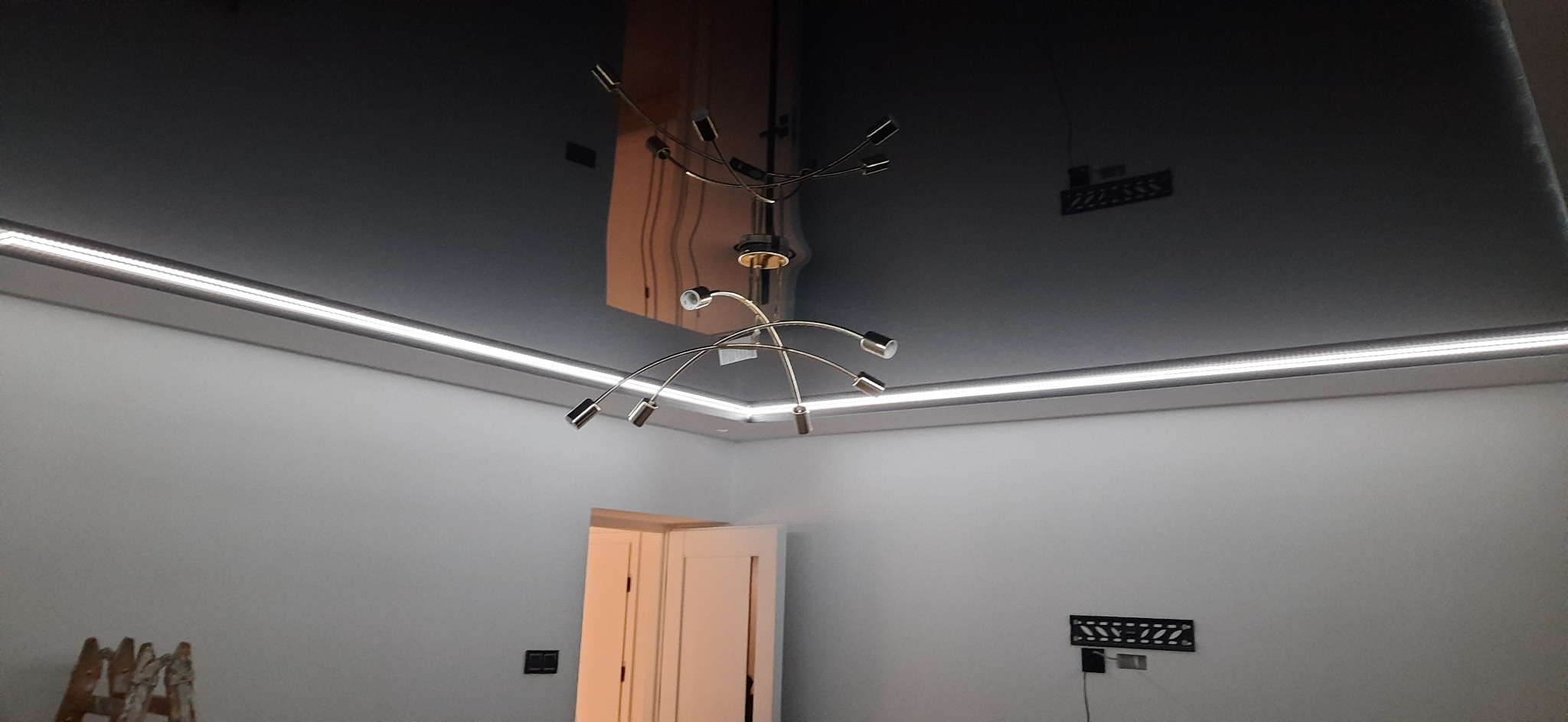 Błyszczący, czarny sufit napinany z oświetleniem LED po obwodzie i nowoczesnym żyrandolem. Widoczne odbicie w suficie. Minimalistyczne wnętrze z białymi ścianami.