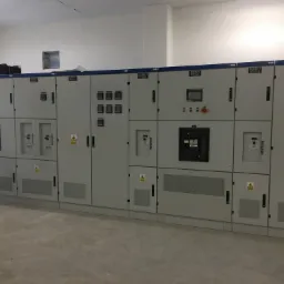 Rozbudowana szafa sterownicza z szarymi panelami i ostrzeżeniami o napięciu, zainstalowana w jasnym, pustym pomieszczeniu z betonową podłogą.