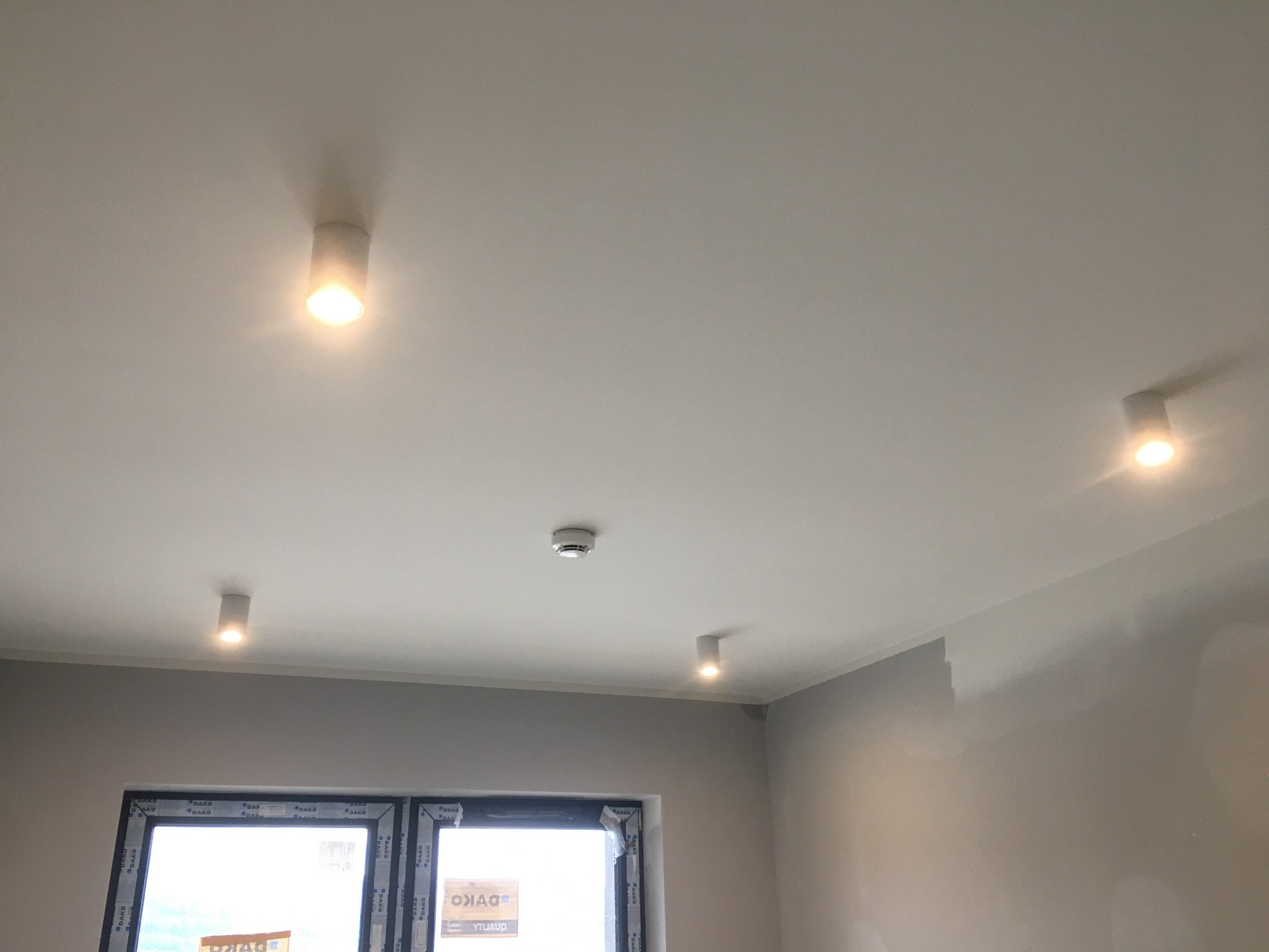 Wnętrze w stanie deweloperskim z zamontowanymi reflektorami punktowymi w suficie i czujnikiem dymu, widoczne okno z naklejkami producenta.