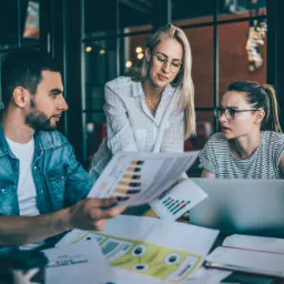 Trzy osoby analizują wykresy i dane finansowe w biurze; blondynka w okularach pochyla się nad dokumentami, mężczyzna w dżinsowej kurtce trzyma arkusz z kolorowymi słupkami, a kobieta w okularach...