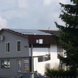 Panele fotowoltaiczne na dachu dwukondygnacyjnego budynku z szarą elewacją, widoczne zza ogrodzenia i drzewa iglastego w słoneczny dzień.