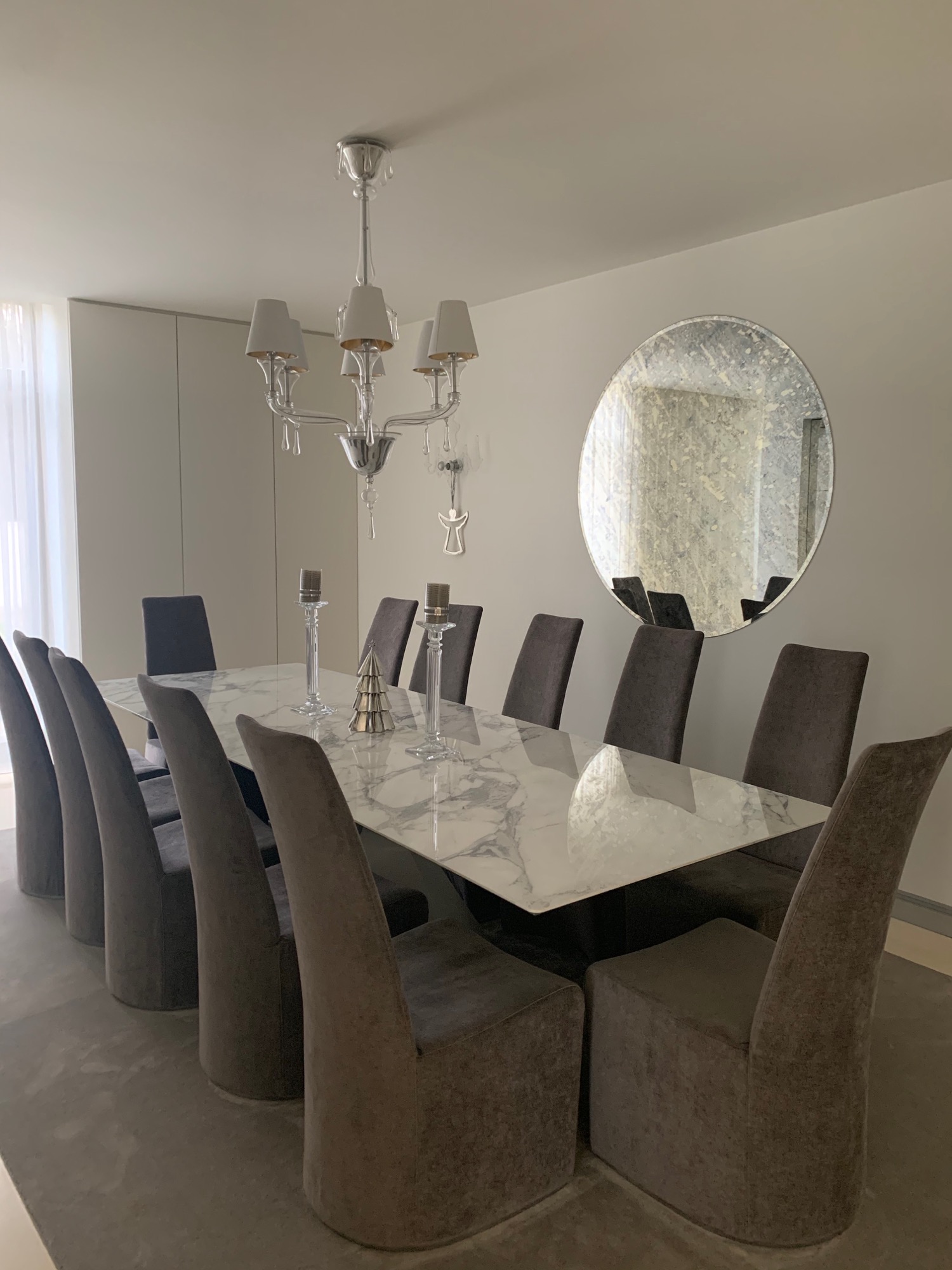 Jadalnia z dużym stołem z marmurowym blatem, obita szarymi krzesłami, ozdobiona srebrnymi świecznikami i dekoracyjną choinką, oświetlona kryształowym żyrandolem, z okrągłym lustrem na ścianie.