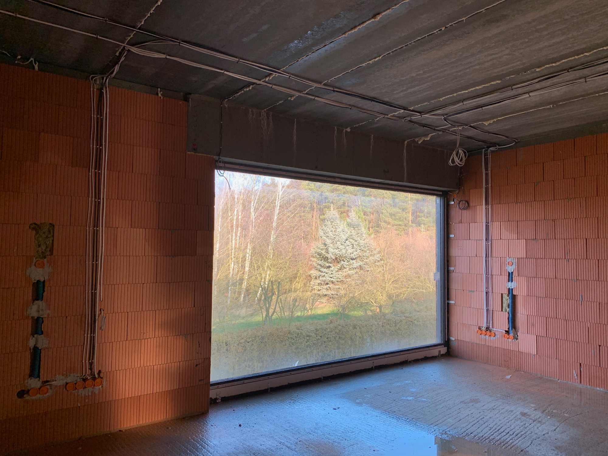 Surowy stan wnętrza z widoczną instalacją elektryczną w ścianach z cegły ceramicznej, kable poprowadzone po suficie, duże okno z widokiem na drzewa w tle.