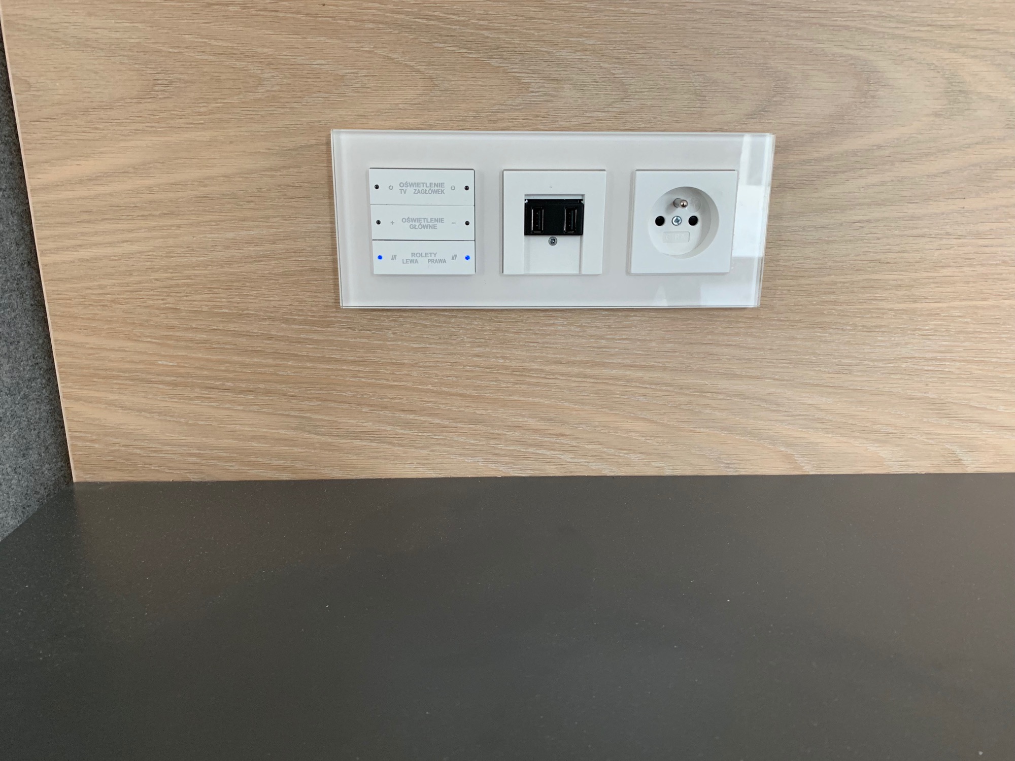 Panel sterowania oświetleniem i roletami z gniazdem elektrycznym i USB, zamontowany na drewnianej ścianie.
