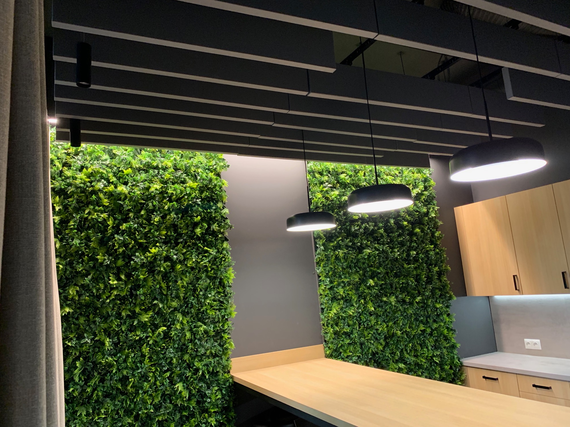 Nowoczesne biuro z zieloną ścianą, drewnianym blatem i oświetleniem punktowym. Sztuczna roślinność na ścianach, minimalistyczne szafki kuchenne w tle.