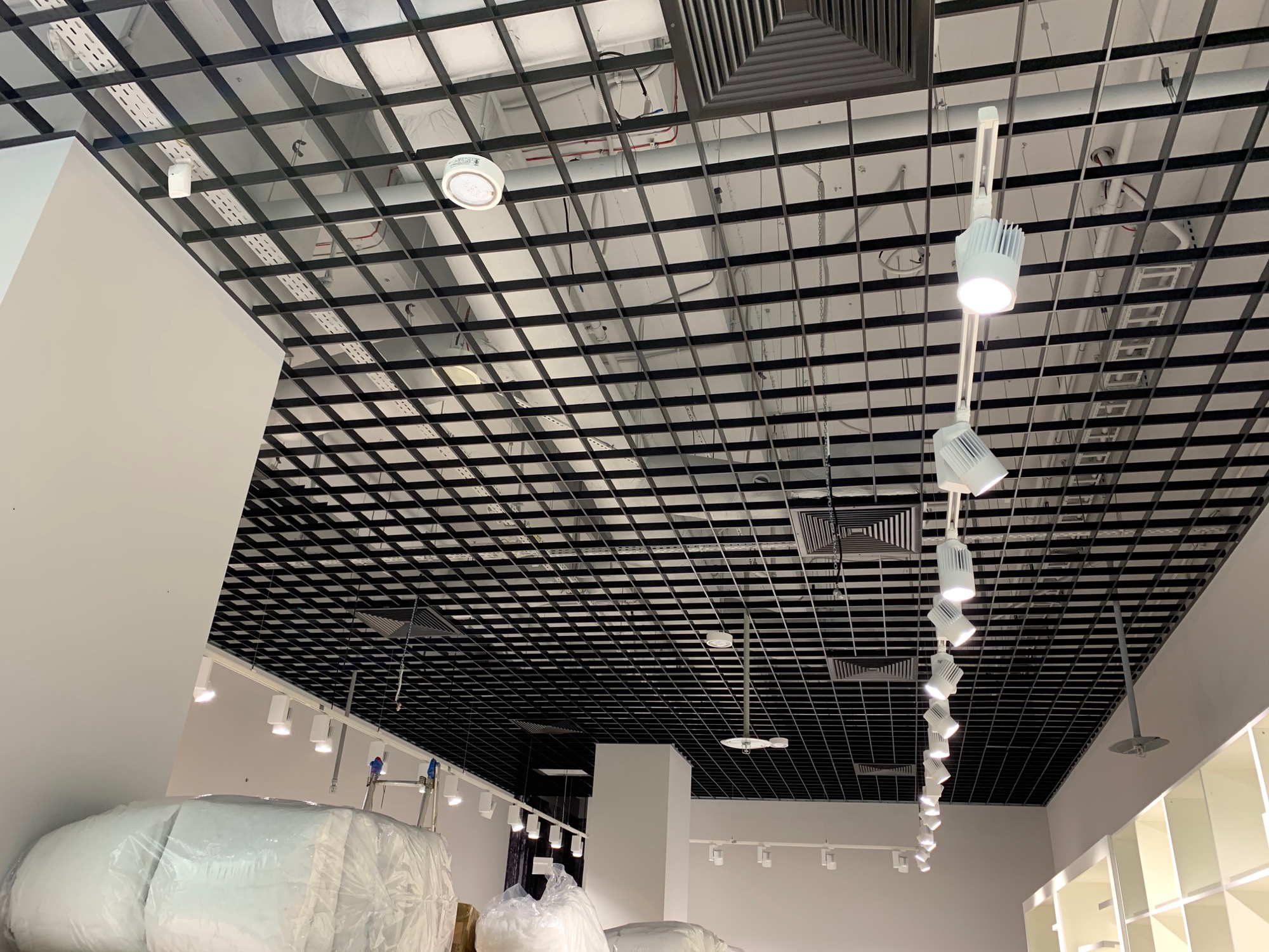 Sufit podwieszany z czarną kratownicą, widoczne białe lampy punktowe, rury wentylacyjne i elementy instalacji elektrycznej w nowym sklepie.