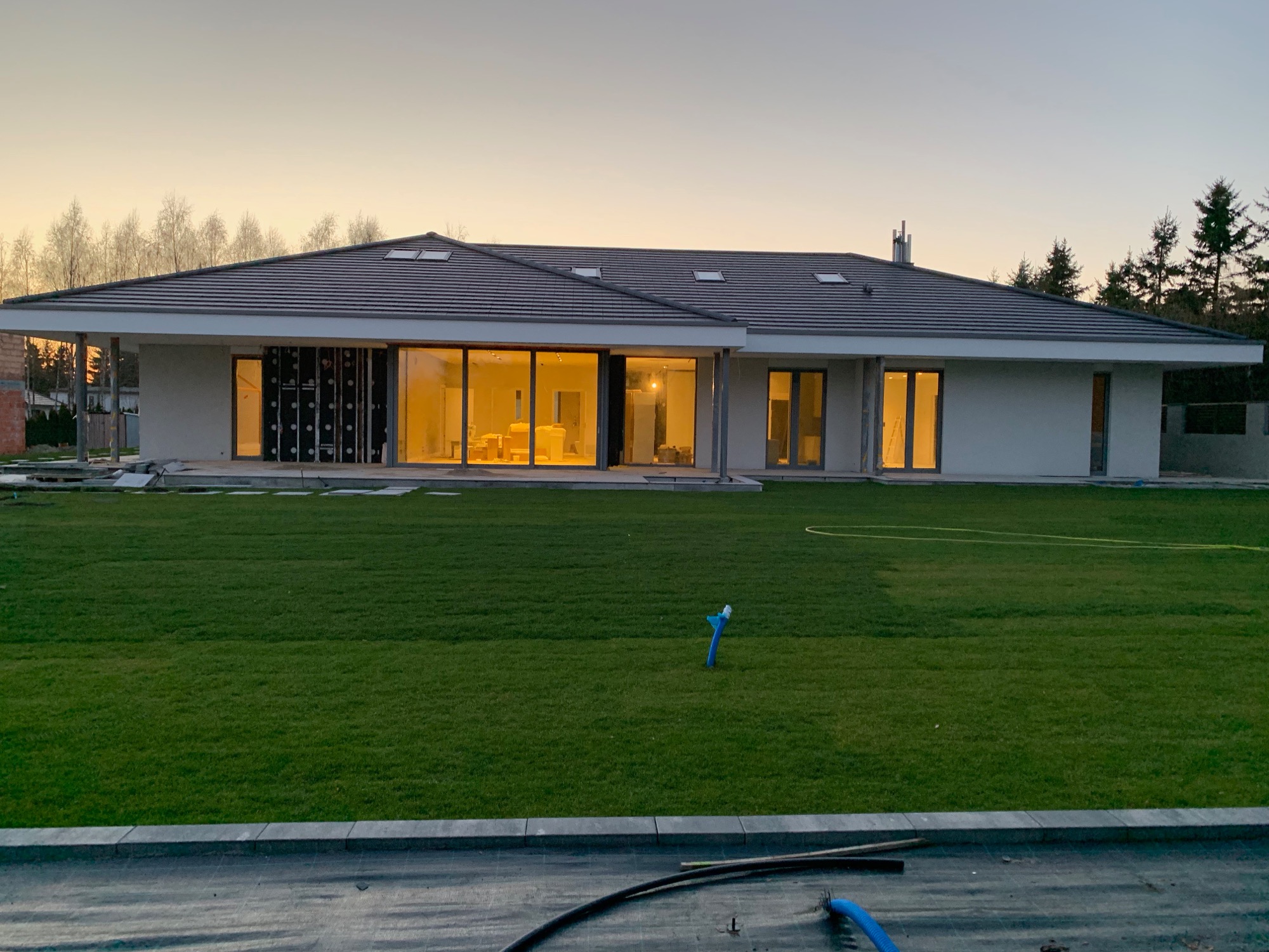 Nowoczesny dom jednorodzinny w trakcie budowy, widoczne duże okna, instalacja elektryczna wewnątrz, zielony trawnik i elementy instalacji na zewnątrz.