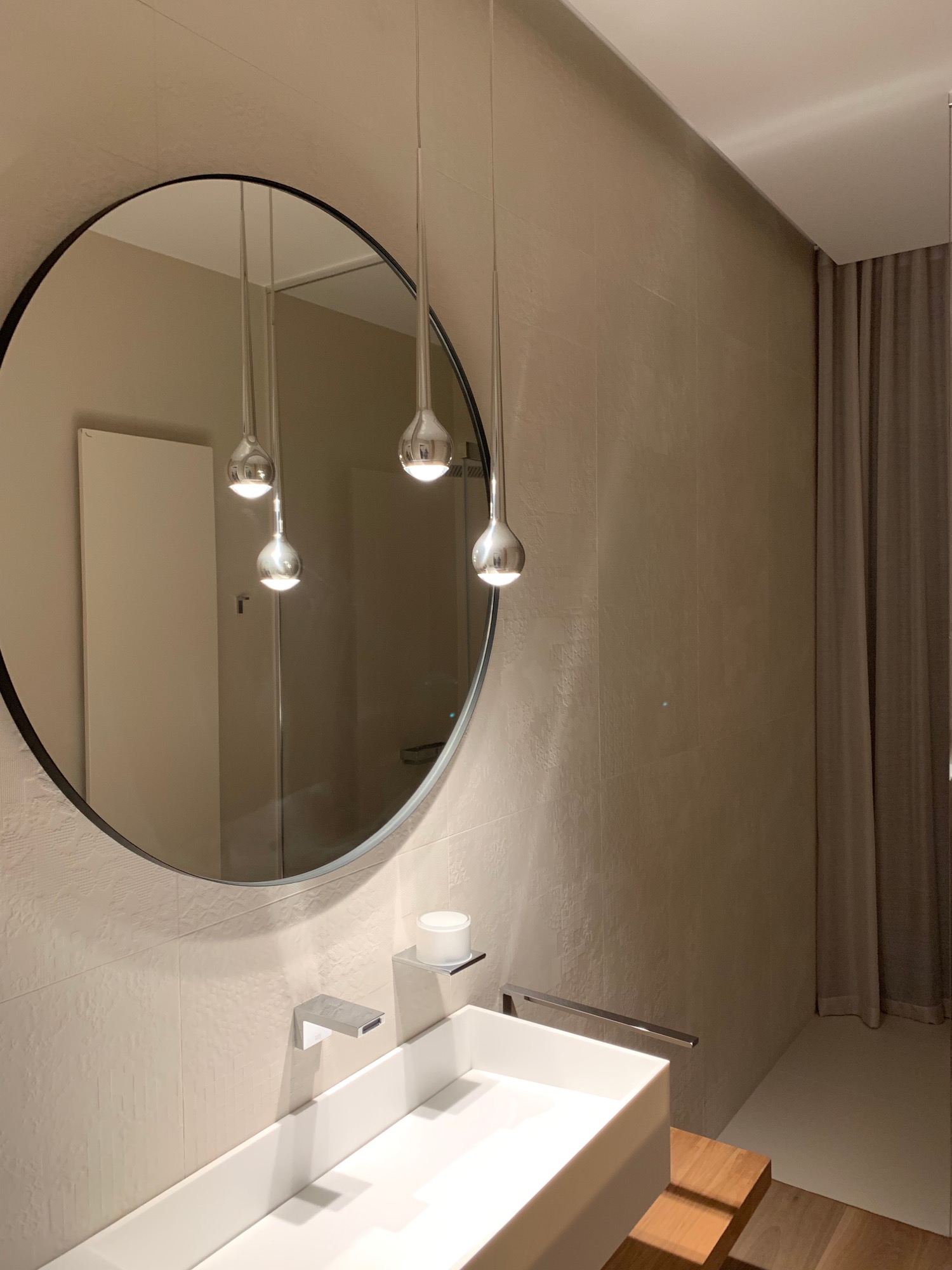 Nowoczesna łazienka z okrągłym lustrem w czarnej ramie, wiszącymi lampami w kształcie kropli i białą, prostokątną umywalką na drewnianym blacie. W tle widoczny grzejnik i kabina prysznicowa.