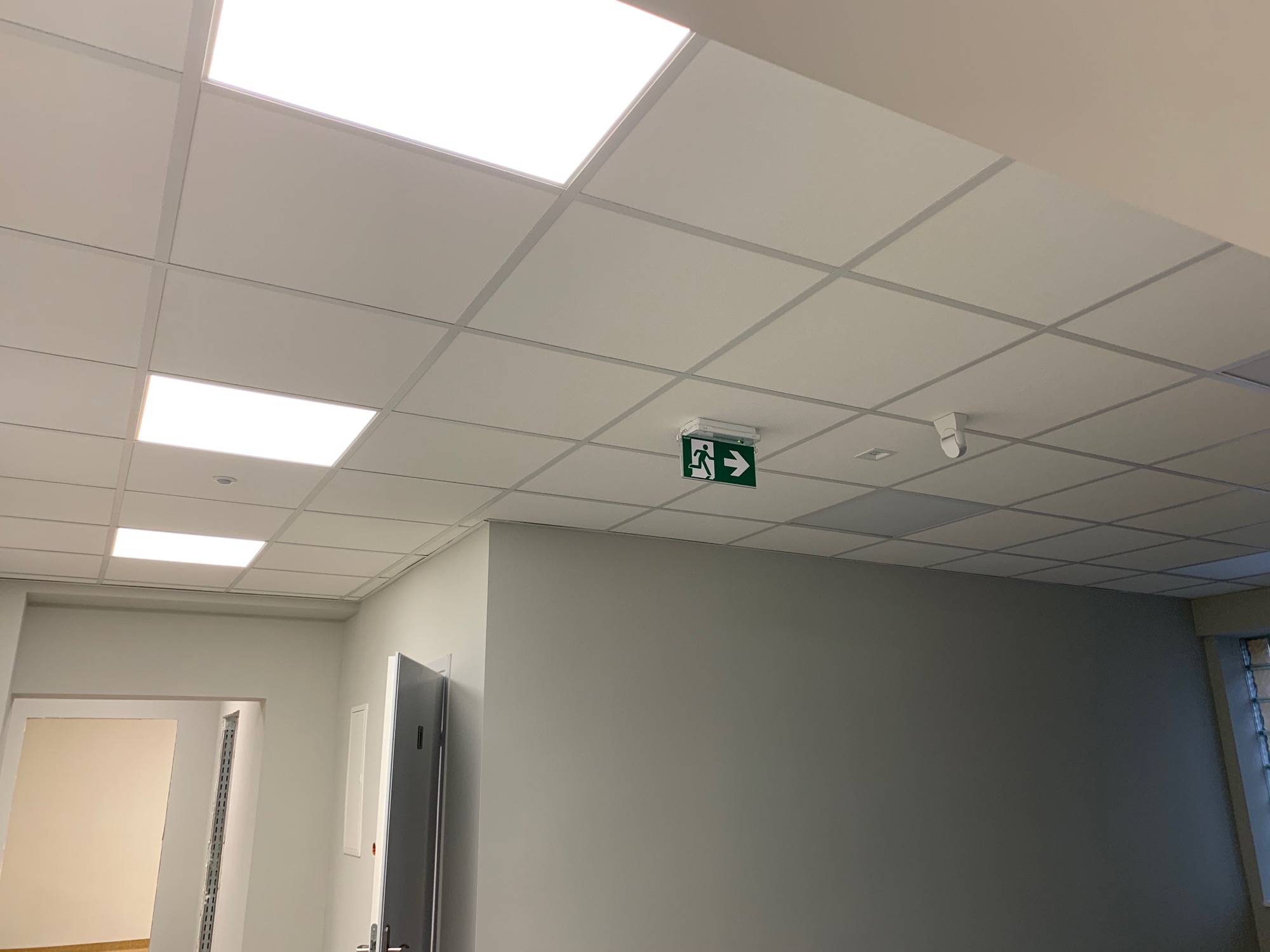 Sufit podwieszany z rastrem, oświetleniem LED i zielonym znakiem ewakuacyjnym wskazującym kierunek w prawo, zamontowanym w korytarzu biurowym.