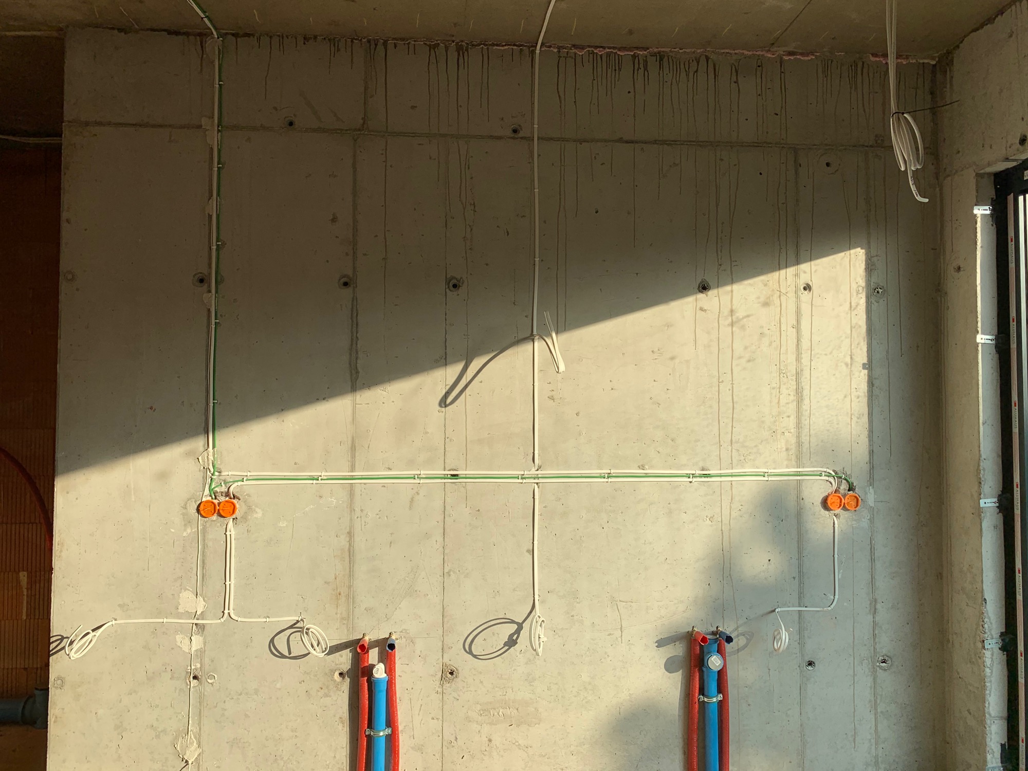 Surowa betonowa ściana z widocznymi przewodami elektrycznymi w peszlach i puszkach instalacyjnych, częściowo oświetlona promieniami słonecznymi, instalacja wodna z rurami w kolorze niebieskim...