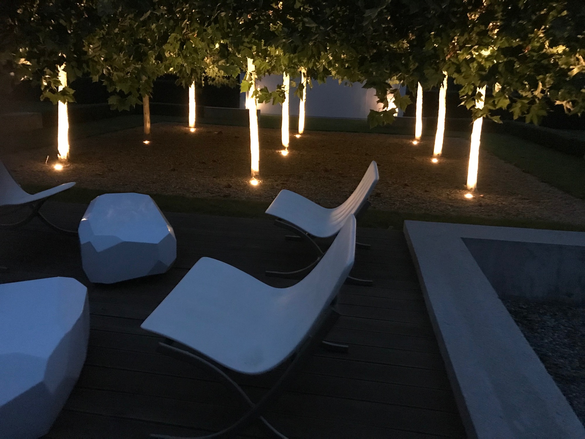 Oświetlenie dekoracyjne ogrodu z białymi, nowoczesnymi meblami ogrodowymi i drzewami podświetlonymi od dołu ciepłym światłem, tworzącymi nastrojową atmosferę.
