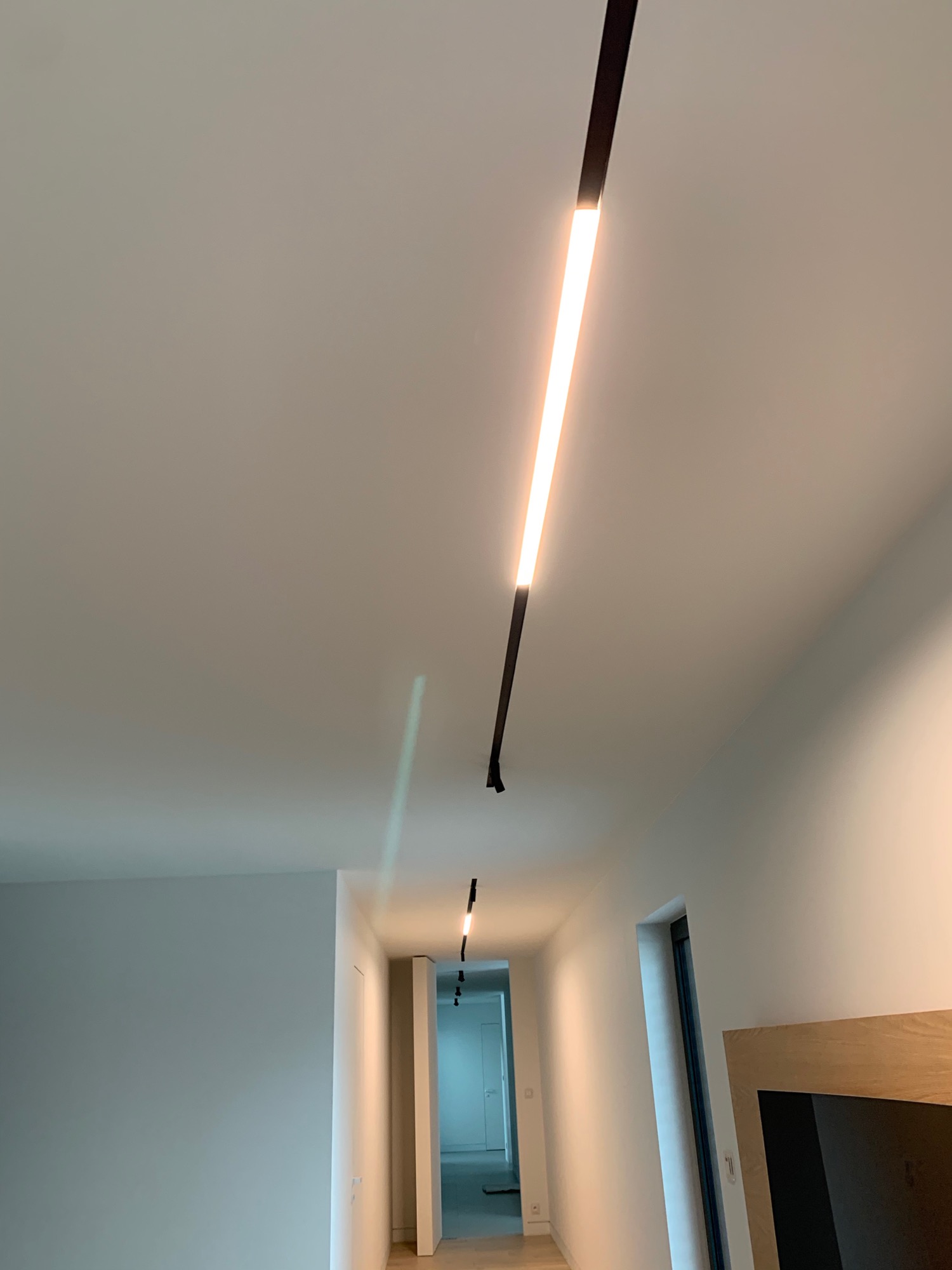 Długi, smukły panel LED w czarnej obudowie, zamontowany na suficie w nowoczesnym korytarzu, z widocznymi kolejnymi panelami w oddali, oświetlającymi przejście.