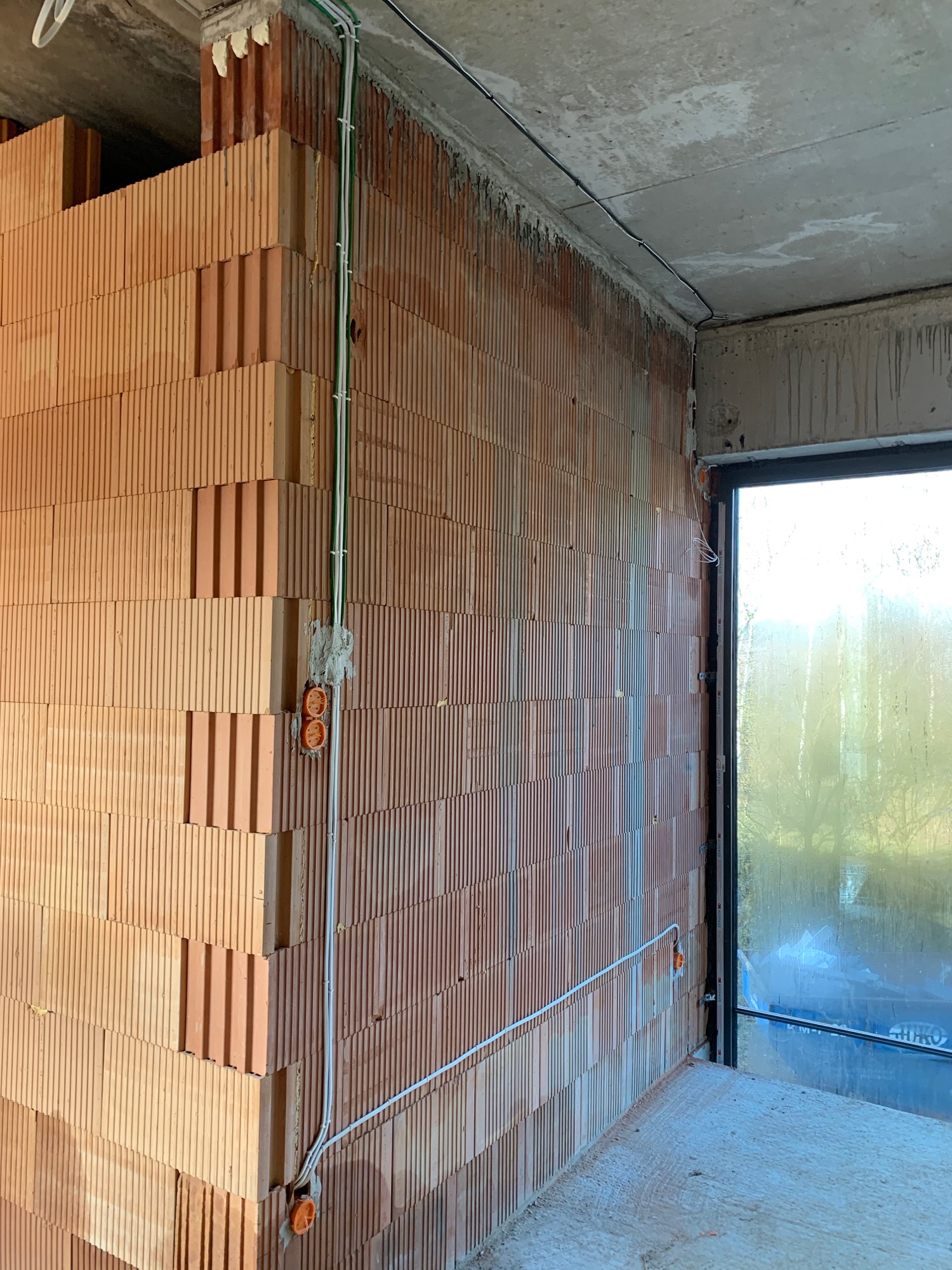 Wnętrze w stanie surowym: ściany z czerwonej cegły dziurawki, puszki elektryczne natynkowe pomarańczowe, przewody elektryczne w białych rurkach PCV, okno z widokiem na zewnątrz.