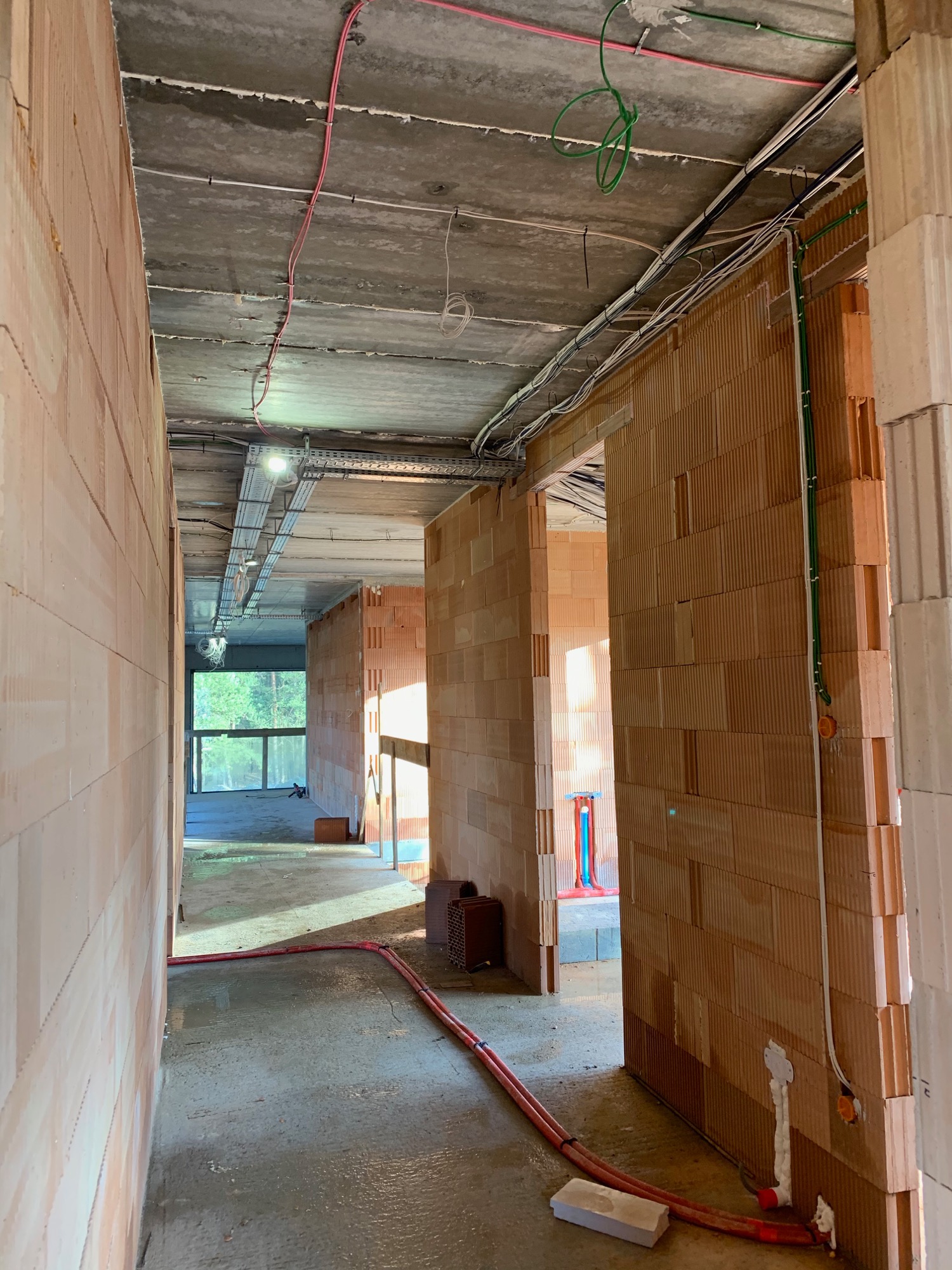 Wnętrze surowego budynku w trakcie prac instalacyjnych: widoczne kable elektryczne prowadzone pod sufitem i pionowo po ścianach z cegły, podłoga w stanie surowym, w tle okno z widokiem na zieleń.
