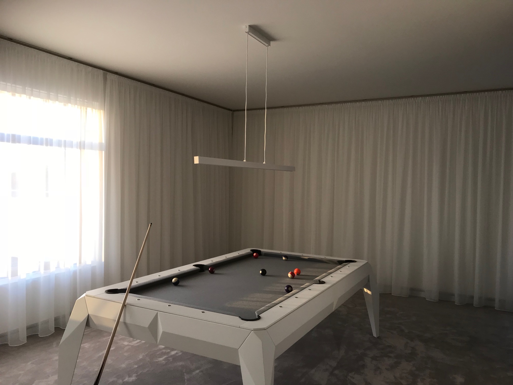Nowoczesny stół bilardowy w jasnym pomieszczeniu z białymi zasłonami i minimalistyczną lampą wiszącą, kij bilardowy oparty o stół.