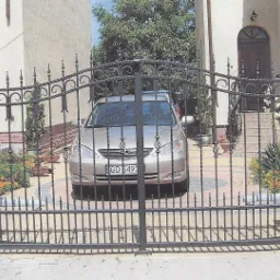 Kuta brama wjazdowa z dekoracyjnymi elementami, z widocznym samochodem osobowym za nią i domem w tle.