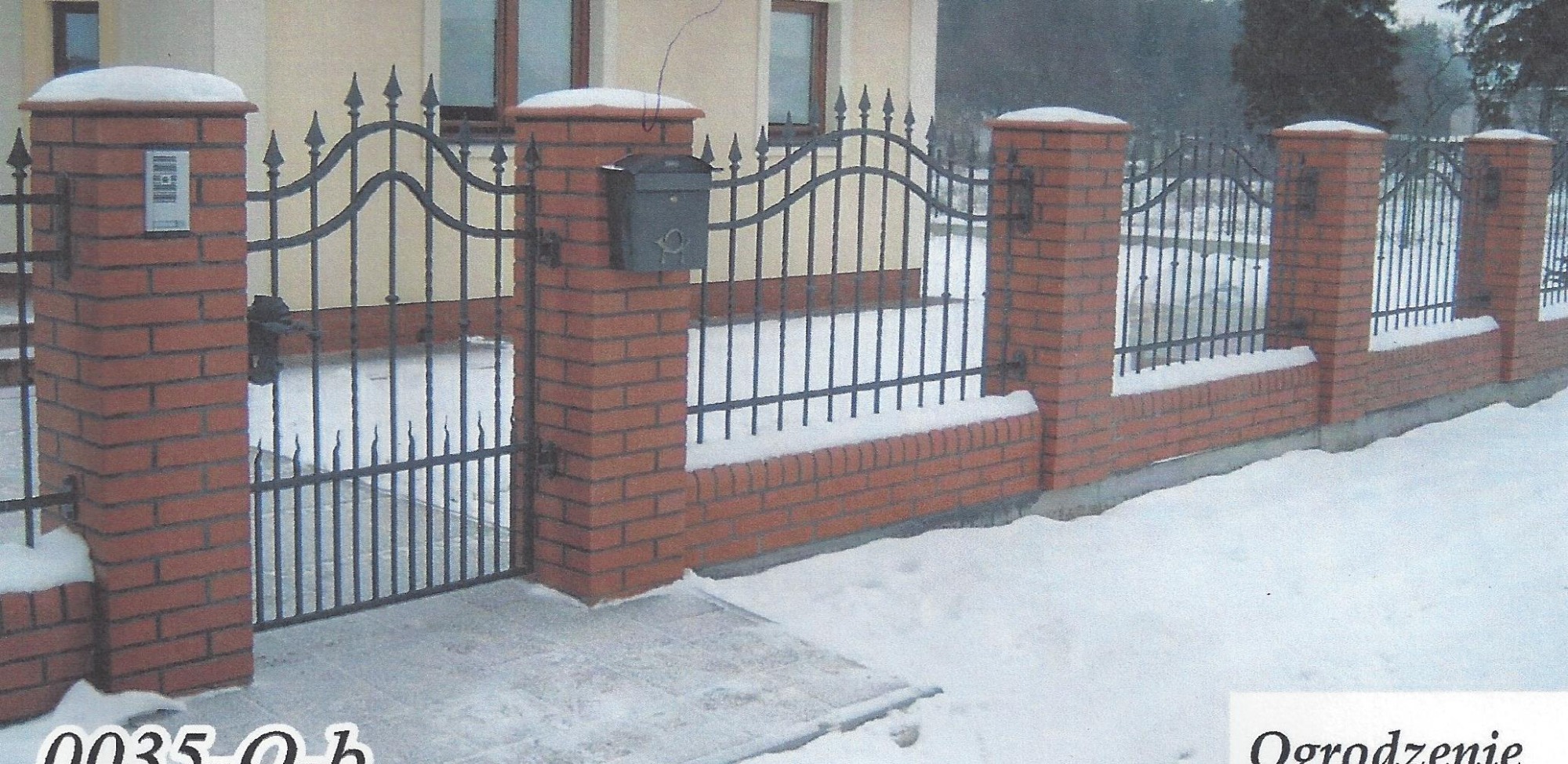 Kuta furtka z czarnego metalu pomiędzy murowanymi słupkami z czerwonej cegły, część ogrodzenia w zimowej scenerii z pokrywą śnieżną na elementach konstrukcyjnych i skrzynką pocztową.