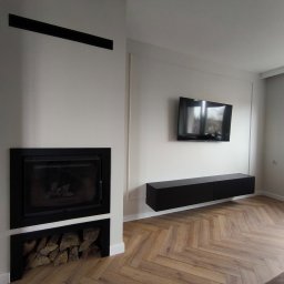 DAWID ŚWIDER DAV HOUSE - Minimalistyczny salon z kominkiem w czarnej obudowie i drewnianą podłogą ułożoną w jodełkę. Na ścianie telewizor i podwieszana szafka RTV.