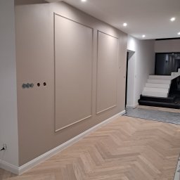 DAWID ŚWIDER DAV HOUSE - Beżowa ściana z ozdobnymi ramami i instalacją elektryczną, parkiet w jodełkę, białe listwy przypodłogowe i schody w tle. Nowoczesne wykończenie.