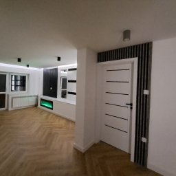 DAWID ŚWIDER DAV HOUSE - Salon po remoncie: parkiet w jodełkę, białe ściany, nowoczesne oświetlenie i drzwi z czarnymi akcentami. Widoczny fragment zabudowy z podświetleniem.