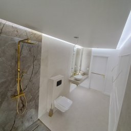 DAWID ŚWIDER DAV HOUSE - Wykończona łazienka w Nowym Targu: marmurowa ściana z złotą armaturą, minimalistyczna toaleta i umywalka na tle białych ścian z subtelnym oświetleniem.