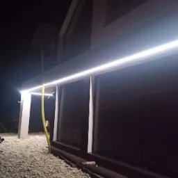 Nocne ujęcie elewacji domu z podświetleniem LED, żwirowe podłoże i widoczny wąż ogrodowy.