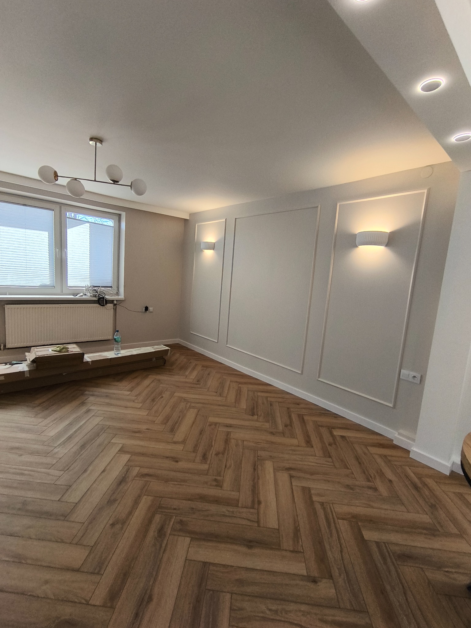 Jasne wnętrze w trakcie wykończenia: parkiet w jodełkę, białe ściany z dekoracyjnymi listwami i nowoczesne oświetlenie. Widoczne materiały budowlane.