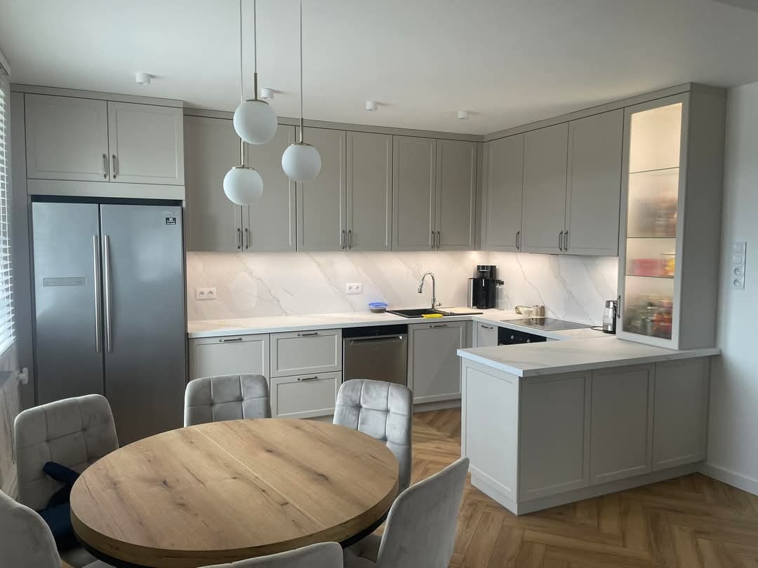 Jasna, nowoczesna kuchnia z szarymi szafkami, marmurowym blatem i drewnianym stołem. Trzy okrągłe lampy wiszące nad stołem. Parkiet w jodełkę na podłodze.