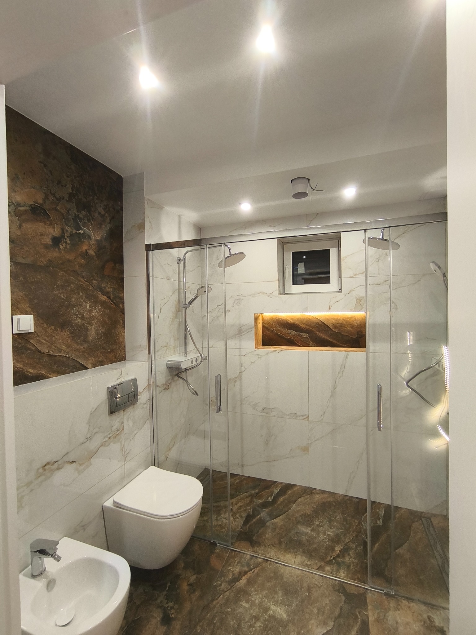 Nowoczesna łazienka z kabiną prysznicową typu walk-in, marmurowymi płytkami i akcentem w postaci podświetlanej wnęki. Bidet i toaleta podwieszana.