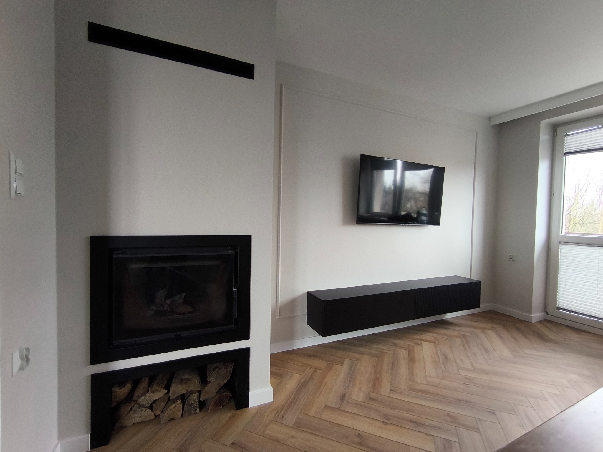 Minimalistyczny salon z kominkiem w czarnej obudowie i drewnianą podłogą ułożoną w jodełkę. Na ścianie telewizor i podwieszana szafka RTV.